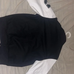 Svart och vit jacka från American College - Snygg svart och vit jacka från American College med kontrasterande ärmar och broderad logga på ärmen. Jackan har knappar framtill och praktiska fickor med vita detaljer. Perfekt för en sportig look!