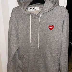 Grå hoodie från Comme des Garçons Play - Säljer en stilren grå hoodie från Comme des Garçons Play med det ikoniska röda hjärtat på bröstet. Tröjan har en justerbar huva med vita snören och en praktisk magficka. Perfekt för en avslappnad look. Storlek XL men passar L bra. Prutbar.