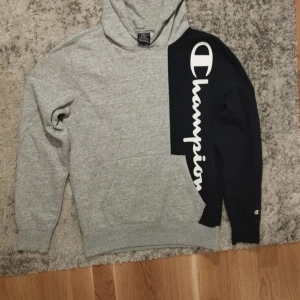 Grå och svart hoodie från Champion - Säljer en cool hoodie från Champion i grått och svart med stor logga på bröstet. Tröjan har en klassisk huva och en stor ficka framtill. Perfekt för en avslappnad stil.