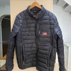 Mörkblå quiltad jacka från Tuxer - Säljer en mörkblå quiltad jacka från Tuxer med dragkedja och en liten bröstficka med flaggdetalj. Jackan har en hög krage och är perfekt för kyligare dagar. Passar bra för både stad och natur.