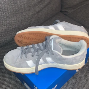 Grå Adidas Campus sneakers - Snygga grå Adidas Campus sneakers med vita detaljer och klassisk gummisula. Perfekta för en avslappnad stil. Skorna har snörning och är tillverkade i mocka för en stilren look.