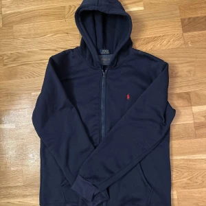 Mörkblå hoodie från Polo Ralph Lauren - Säljer en stilren mörkblå hoodie från Polo Ralph Lauren med dragkedja och den ikoniska röda loggan på bröstet. Perfekt för en avslappnad look.