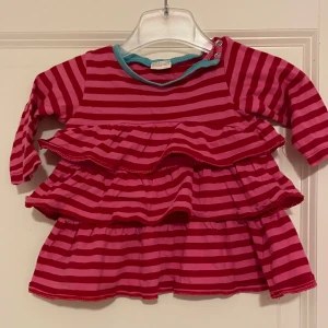 Röd- och rosa randig babyklänning från Lindex storlek 68 - Söt babyklänning från Lindexi röd- och rosa randigt mönster. Klänningen har volanger och långa ärmar med en turkos kant vid halsen. Perfekt för små fashionistas!. Storlek 68.