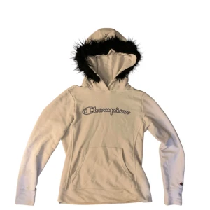 Vit hoodie med svart päls - Snygg vit hoodie från Champion med svart pälsdetalj runt huvan. Den har en klassisk känguruficka framtill och långa ärmar. Perfekt för en stilren och bekväm look. Ställ gärna frågor!