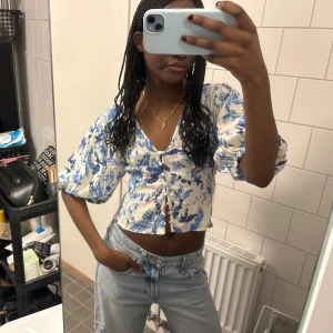 Blommig croppad blus - Säljer en söt blommig blus i blått och vitt med puffiga trekvartsärmar och v-ringning. Blusen är croppad och har knappar framtill, perfekt för sommaren. Använt endast 2 ggr🌸