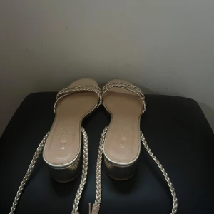 Guldiga sandaler med flätad rem - Snygga guldiga sandaler med flätade remmar och låg klack. Perfekta för en somrig look med en touch av glamour. De har en fyrkantig tå och en elegant design som passar till många tillfällen.