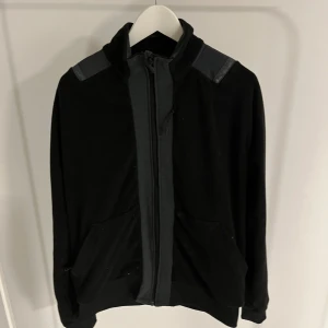 Svart jacka med dragkedja - Snygg svart jacka med dragkedja och detaljer på axlarna. Perfekt för en stilren look. Jackan har långa ärmar och praktiska fickor framtill.