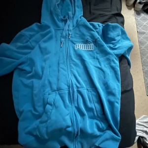 Blå hoodie från Puma - Säljer en blå hoodie från Puma med dragkedja och huva. Tröjan har en broderad Puma-logga på bröstet och är tillverkad i ett mjukt material. Perfekt för en avslappnad stil. Djur finns i hemmet 