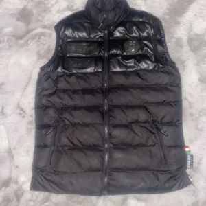 Svart dunväst från Philipp Plein - Snygg svart dunväst från Philipp Plein med glansig finish och dragkedja framtill. Västen har två praktiska fickor med dragkedja och en stilren design med märkets logotyp på bröstet.