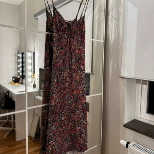 Blommig långklänning med öppen rygg - Snygg långklänning med blommigt mönster i flera färger. Klänningen har smala axelband och en elegant öppen rygg med knytning. Perfekt för sommarens festligheter!