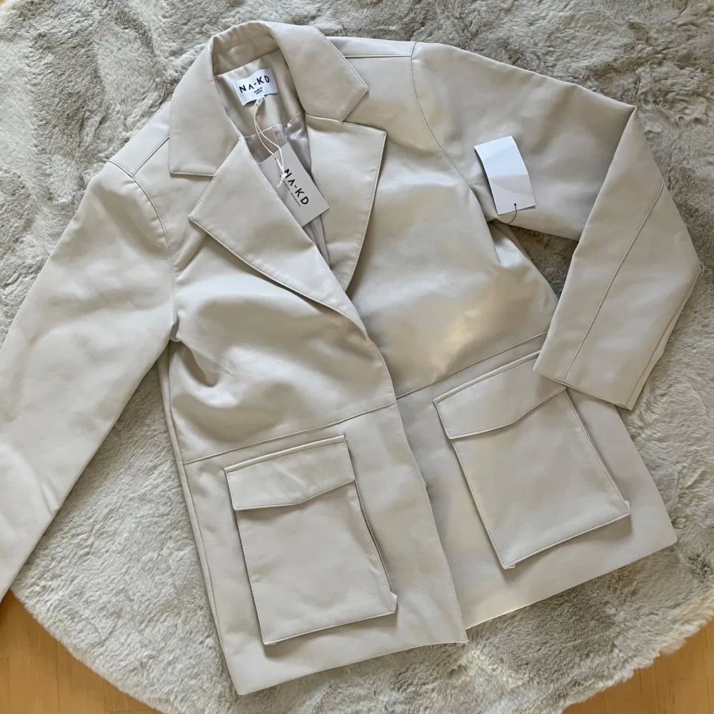 Snygg beige kavaj från NA-KD med klassisk design. Den har långa ärmar och stora fickor framtill. Perfekt för en stilren look.. Takit.