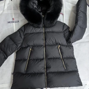 Svart dunjacka från Moncler - Snygg svart dunjacka från Moncler med en fluffig huva och dragkedja framtill. Jackan har långa ärmar och två praktiska fickor med dragkedjor. Perfekt för kyliga dagar.