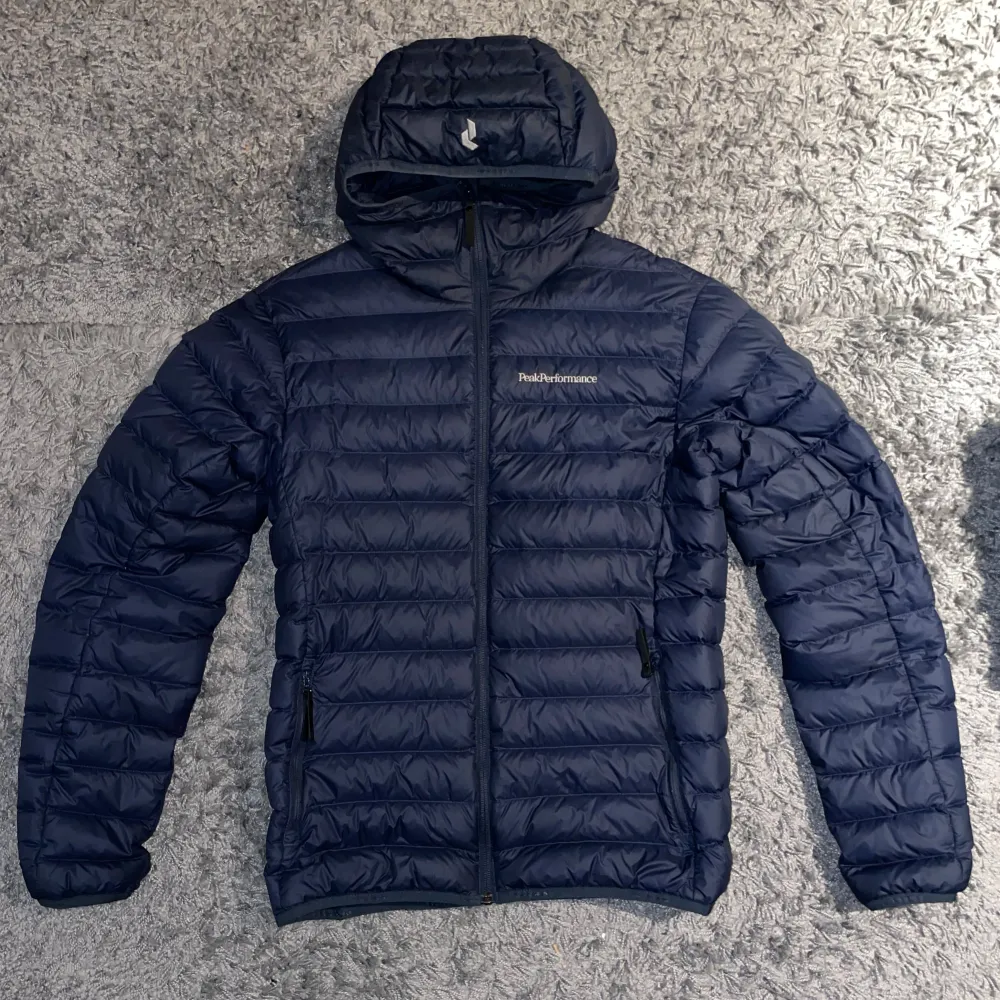 Säljer en snygg blå dunjacka från Peak Performance. Jackan har en quiltad design med dragkedja och huva, perfekt för kyliga dagar. . Takit.