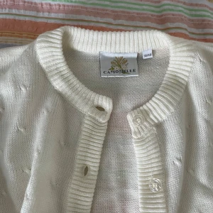 Vit stickad kofta från Camomille - Säljer en superfin vit stickad kofta från Camomille. Den har ett gulligt mönster med små blommor och diamantformer på nedre delen av koftan och ärmarna. Koftan har långa ärmar och knappar hela vägen fram. Perfekt för höst och vår! 🌸