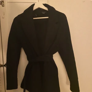 Svart kavaj med bälte - Säljer en snygg svart kort kappa i storlek XS. Den har ett bälte som framhäver midjan. Perfekt material för hösten. Kavajen är långärmad och i nyskick. Den är i jätte bra skick, använd sparsamt. 