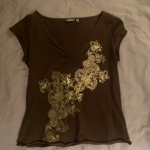 Brun topp med mönster  - Säljer en snygg brun topp!  Toppen har ett vackert mönster i guld och vit på framsidan och en v-ringad hals. Den är kortärmad och gjord av ett mjukt och bekvämt material!