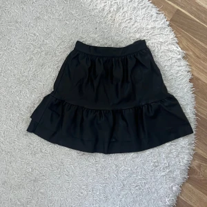 Svart kortkjol med volang - Säljer en superfin svart kortkjol med volangdetaljer. Kjolen är perfekt för både vardag och fest. I skinn