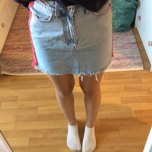 Jeanskjol med fransar - Säljer en snygg jeanskjol i ljusblå denim med fransar längst ner. Kjolen har en normal passform och är perfekt för både vardag och fest. Den har en dragkedja och knappar framtill samt två bakfickor. Superfin att matcha med en t-shirt eller en stickad tröja!