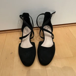 Svarta ballerinaskor med klack och remmar - Säljer ett par snygga svarta ballerinaskor med klack och korsade remmar. Skorna är i mocka och har en bekväm passform. Perfekta för både vardag och fest!