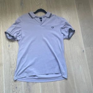 Ljuslila pikétröja från Fred Perry - Säljer en snygg ljuslila pikétröja från Fred Perry. Tröjan har korta ärmar och en klassisk krage med två knappar. Den är i bra skick och perfekt för både vardag och fest. Den har också Fred Perry-loggan broderad på bröstet. Passar perfekt till jeans eller shorts!