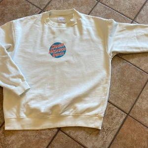 Vit sweatshirt från Vaillent med Santa Monica Beach-tryck - Säljer en superfin vit sweatshirt från Vaillent i storlek S. Den har ett coolt tryck med texten 'Santa Monica Beach' i rött och blått på framsidan. Tröjan är långärmad och gjord av mjuk bomull, perfekt för en avslappnad stil. Passar bra till både jeans och leggings. Perfekt för höst och vår!