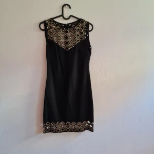 Svart klänning från Nice Istanbul - Säljer en superfin svart klänning från Nice Istanbul i storlek 40. Klänningen är ärmlös och har en tight passform. Den har snygga gulddetaljer och paljetter på både fram- och baksidan, vilket gör den perfekt för fest. Materialet känns som polyester och den har en dragkedja på baksidan för enkel på- och avtagning. Perfekt för en utekväll eller en speciell tillställning!