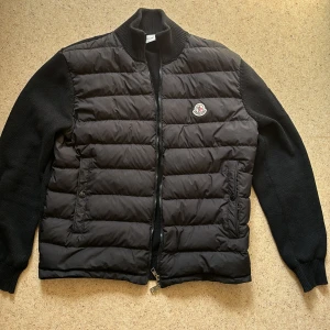 Cardigan Moncler - Säljer en snygg och stilren Moncler-cardigan i svart. Cardiganen har en stickad bas med en fastsydd vadderad väst framtill, vilket ger både värme och ett modernt utseende. Den är försedd med Monclers ikoniska logotyp på bröstet. Perfekt för både 