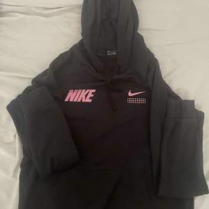 Säljer en svart hoodie från Nike i storlek XS. Den har en stor rosa Nike-logga på bröstet och en liten vit Nike-swoosh bredvid. Hoodien är supermjuk och perfekt för höst och vinter. Den har en stor ficka på magen och en justerbar huva. Perfekt för både träning och vardag!