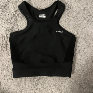 Svart sporttopp från Stronger - Snygg och funktionell svart sporttopp från Stronger. Toppen har en tight passform och är tillverkad i ett syntetmaterial som andas. Perfekt för träning med sitt stilrena och bekväma design.