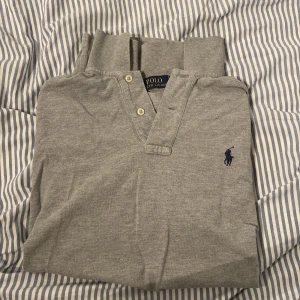 Grå pikétröja från Ralph Lauren - Säljer en klassisk grå pikétröja från Ralph Lauren med den ikoniska loggan broderad i mörkblått på bröstet. Tröjan har en krage och knappar framtill. Perfekt för en stilren look.