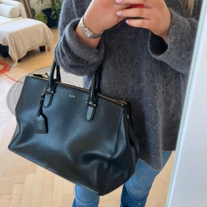 Svart skinnväska från Ralph Lauren - Elegant svart skinnväska från Ralph Lauren med guldfärgade detaljer och dragkedja. Väskan har två handtag och flera fack inuti för praktisk förvaring. Perfekt för en stilren look. Superfint skick!!!