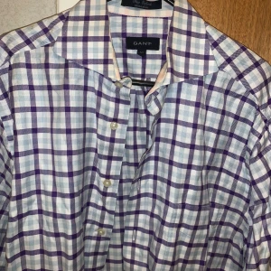 Rutig skjorta från GANT - Säljer en stilren rutig skjorta från GANT i storlek M. Skjortan har ett klassiskt rutmönster i lila och vitt med knappar framtill.