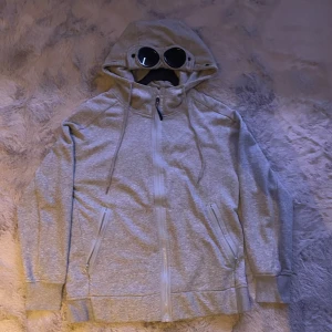 C.P. Company - Säljer en grå huvtröja från C.P. Company med inbyggda glasögon i huvan. Tröjan har dragkedja framtill och två fickor med dragkedjor. Perfekt för en unik och stilren look.