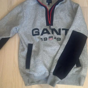 Grå sweatshirt från GANT - Säljer en grå sweatshirt från GANT med svart och röd dragkedja. Tröjan har svarta detaljer på ärmarna och ett tryck med texten 'GANT 1949' på framsidan. Perfekt för en avslappnad stil. Säljer åt min bror då den inte passade honom, men den är i nyskick. 