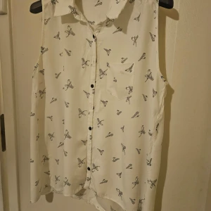 Vit ärmlös blus från Dorothy Perkins - Säljer en vit ärmlös blus från Dorothy Perkins med ett svart fjärilsmönster. Blusen har en klassisk krage och knappar framtill. Perfekt för en stilren och luftig look.