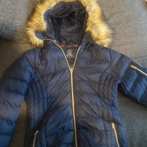 Mörkblå quiltad jacka med fuskpäls - Säljer en mörkblå quiltad jacka från Geographical Norway med fuskpäls på huvan. Jackan har dragkedja och flera praktiska fickor. Perfekt för kyliga dagar!