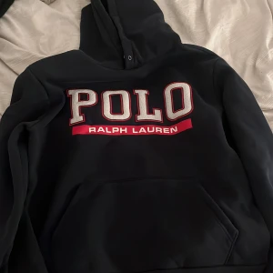 Svart hoodie från Ralph Lauren - Säljer en svart hoodie från Ralph Lauren med stor POLO-logga i rött och vitt på framsidan. Tröjan har en klassisk känguruficka och en bekväm huva. Perfekt för en avslappnad stil.