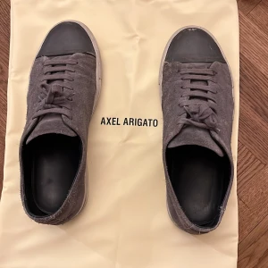 Grå sneakers från Axel Arigato - Snygga grå sneakers från Axel Arigato med svart tåhätta och snörning. Skorna har en stilren design med en vit sula som ger en modern look. Perfekta för en avslappnad stil. Pris kan självklart diskuteras!