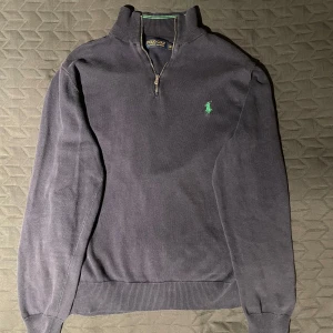 Polo Ralph Lauren tröja - Snygg mörkblå tröja från Polo Ralph Lauren med halv dragkedja och klassisk krage. Den har ett grönt broderat logotyp på bröstet och ribbade muddar vid ärmslut och nederkant.
