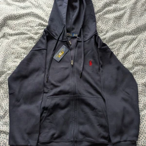 Royal blue hoodie från Ralph Lauren - Säljer en stilren mörkblå hoodie från Ralph Lauren med dragkedja och röd broderad logga på bröstet. Perfekt för en avslappnad look med justerbar huva och fickor framtill. Passar både till vardags och chill. (Hoodien passar bra också för size S.)
