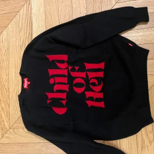 Svart stickad  tröja från Supreme med röd text - Snygg svart tröja från Supreme med röd text 'Child of Hell' på framsidan. Perfekt för en cool och avslappnad stil. Tröjan har långa ärmar och en klassisk rund halsringning.