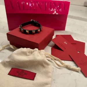 Svart armband med nitar från Valentino - Snyggt svart armband från Valentino dekorerat med guldiga pyramidformade nitar. Perfekt accessoar för att ge en edgy touch till din outfit. Levereras i originalförpackning med tillhörande påse och kort.