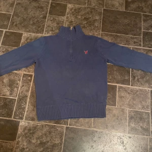 Blå tröja med dragkedja från American Eagle - Säljer en blå tröja från American Eagle med dragkedja vid halsen och ett rött broderat emblem på bröstet. Tröjan har långa ärmar och ribbade muddar vid ärmslut och nederkant. Perfekt för kyliga dagar!