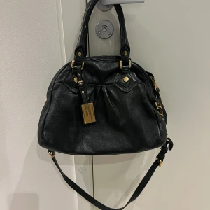 Marc Jacobs väska  - Snygg svart axelväska i skinn med gulddetaljer och dragkedja. Väskan har ett mönstrat innerfoder med bokstäver och justerbar axelrem. Perfekt för att bära med sig det nödvändigaste med stil💞