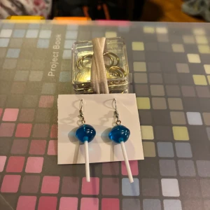 Blå örhängen i form av klubbor - Unika örhängen som ser ut som blå klubbor. Perfekta för att ge en lekfull touch till din stil. De hänger från silverfärgade krokar och är ett roligt tillskott till smyckesamlingen.