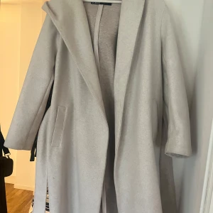 Beige kappa med huva från Zara - Snygg beige kappa med huva och knytskärp i midjan. Perfekt för kyliga dagar med sin långa design och praktiska fickor. En stilren och tidlös jacka för alla tillfällen.