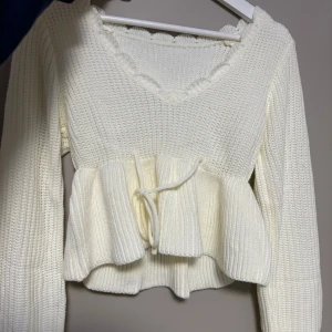 Vit stickad tröja med volangdetalj - Säljer en söt vit stickad tröja med volangdetalj och knytband i midjan. Tröjan har en v-ringad hals och långa ärmar. Den är aldrig använd för den var fel storlek 
