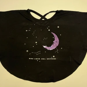 Svart topp med paljettdetaljer - Snygg svart topp med ett unikt paljettmönster i form av en måne och stjärnor på framsidan. Toppen har en korsad ryggdetalj och texten 'Mon Cher Bel Univers'. Perfekt för en stjärnklar kväll!