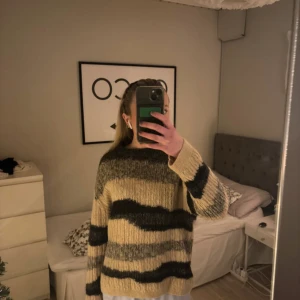 Randig stickad tröja - Säljer en mysig stickad tröja med breda ränder i beige, svart och grått. Tröjan har långa ärmar och en avslappnad passform, perfekt för kyliga dagar. Passar bra till jeans eller leggings för en avslappnad look.