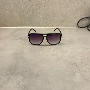 Svarta solglasögon med lila tonade linser - Snygga svarta solglasögon med en klassisk design och lila tonade linser. Bågarna har en stilren och modern look med en tunn metallbro över näsan. Perfekta för att ge en cool touch till din stil. Tänk och ha dom här till en sommardag.